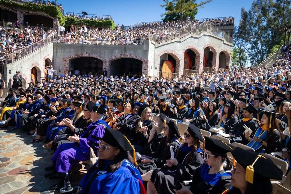 Graduates in caps, gowns and hoods applauding in a sunlit outdoor amphitheater filled with families and guests at commencement.