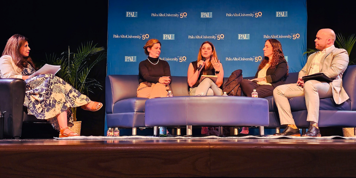 Panel discussion on stage in front of a Palo Alto University 50th anniversary backdrop, with a moderator and four panelists seated, one speaking into a microphone.